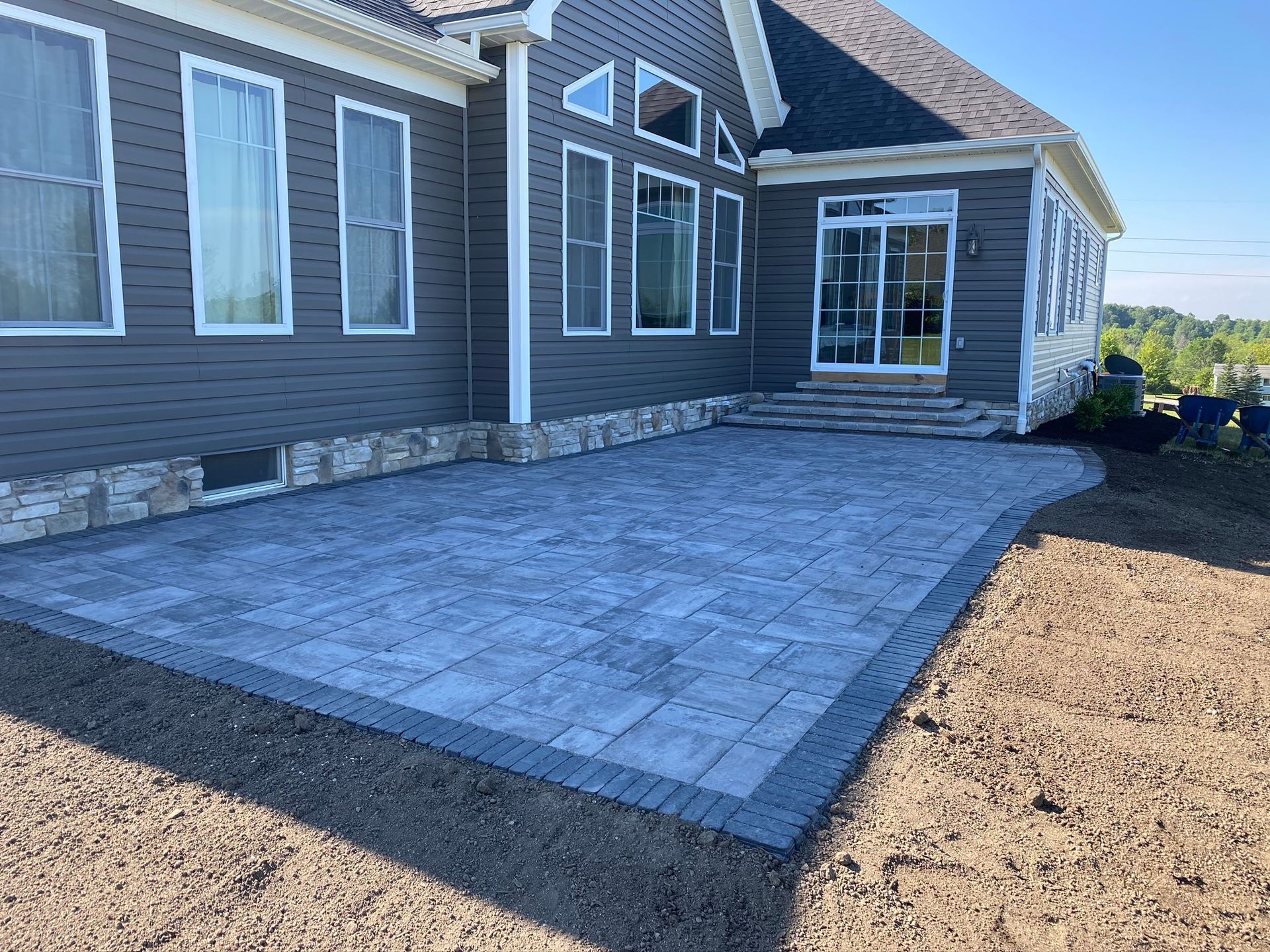 Stone patio beside a gray house with sliding glass doors and windows, bordered by bare dirt.