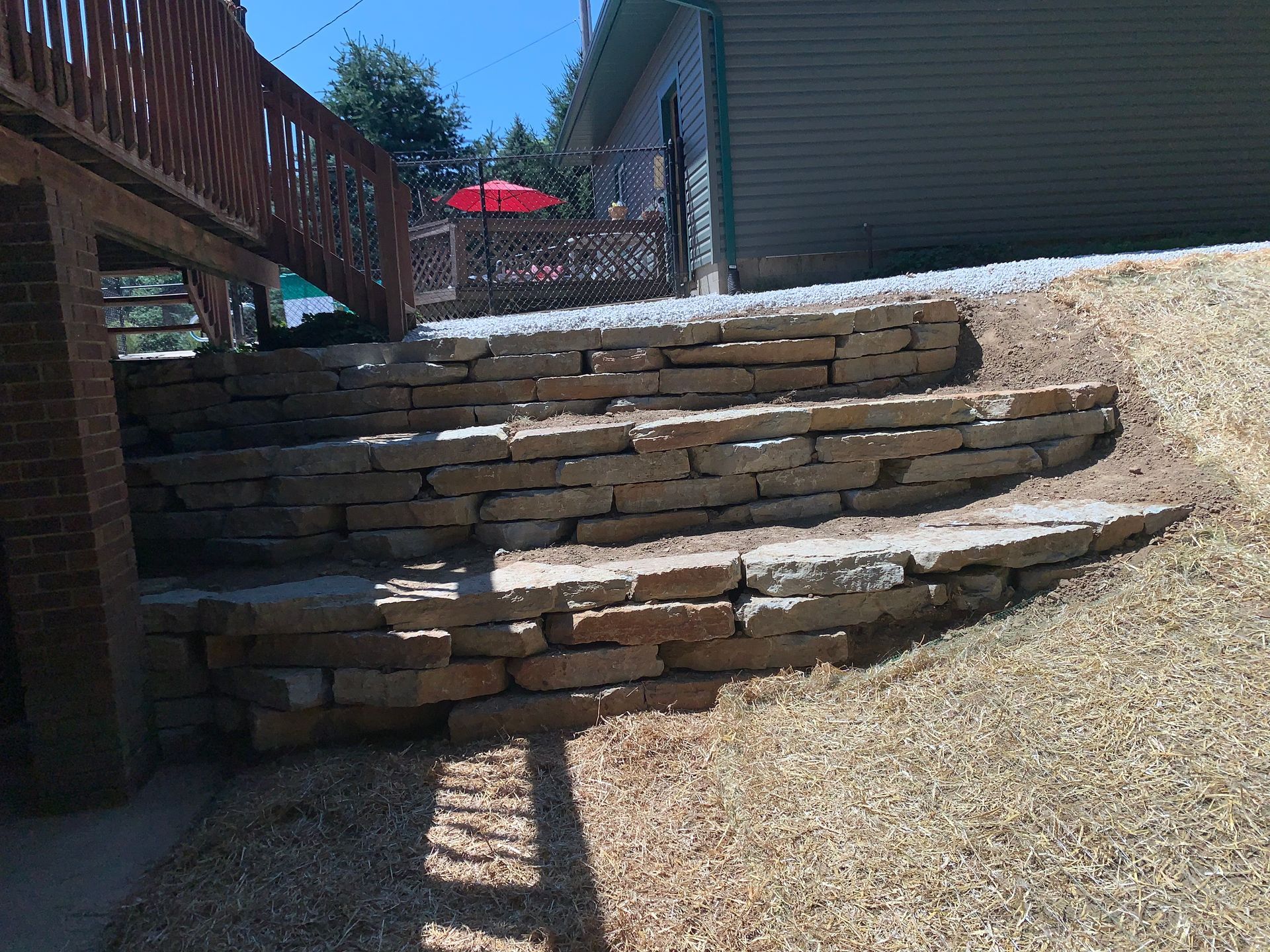 Stone retaining wall and steps in a backyard with a deck and red umbrella.