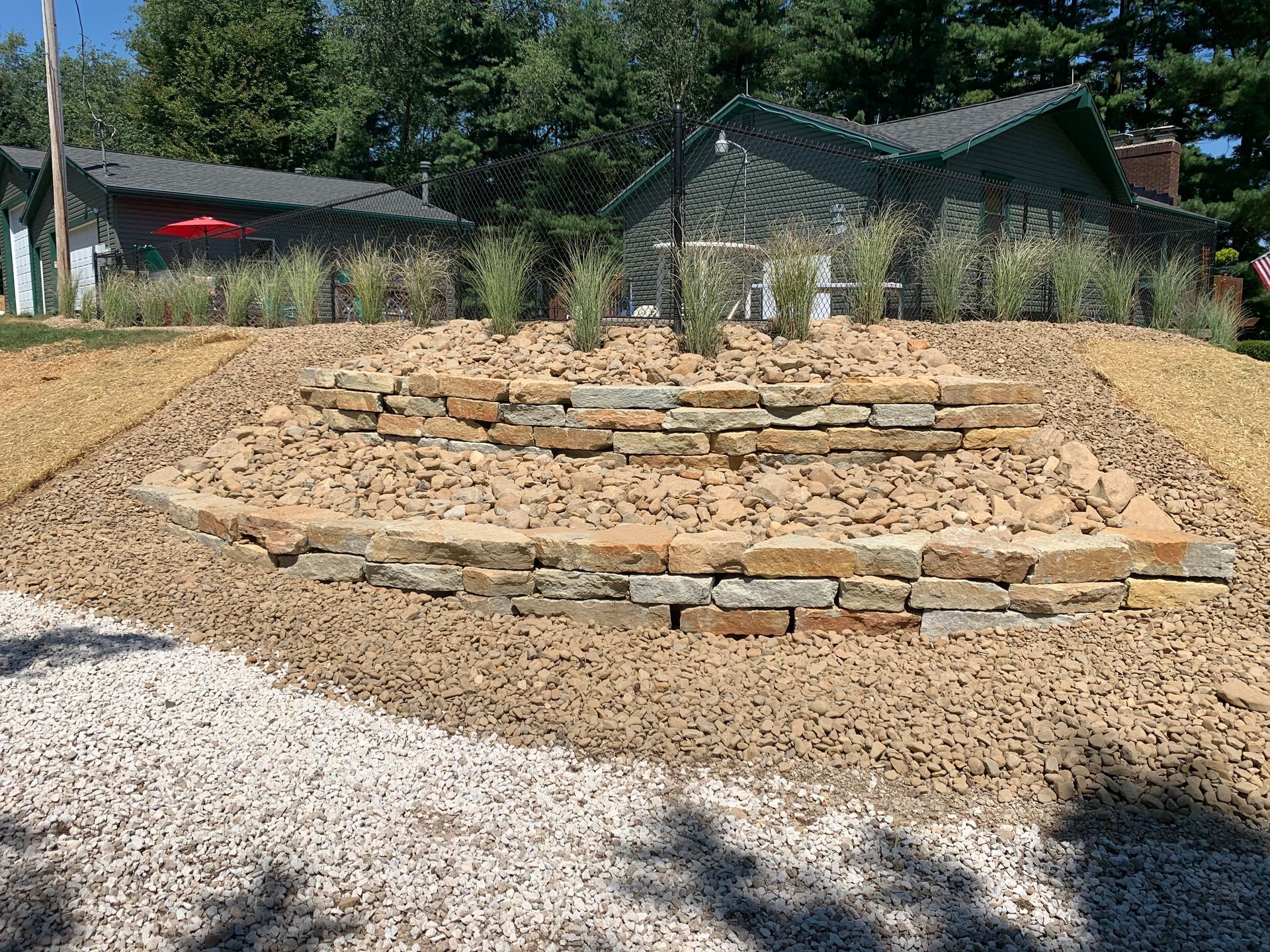 Circular stone garden bed in a sunny yard, with gravel foreground and trees and a greenhouse in back.