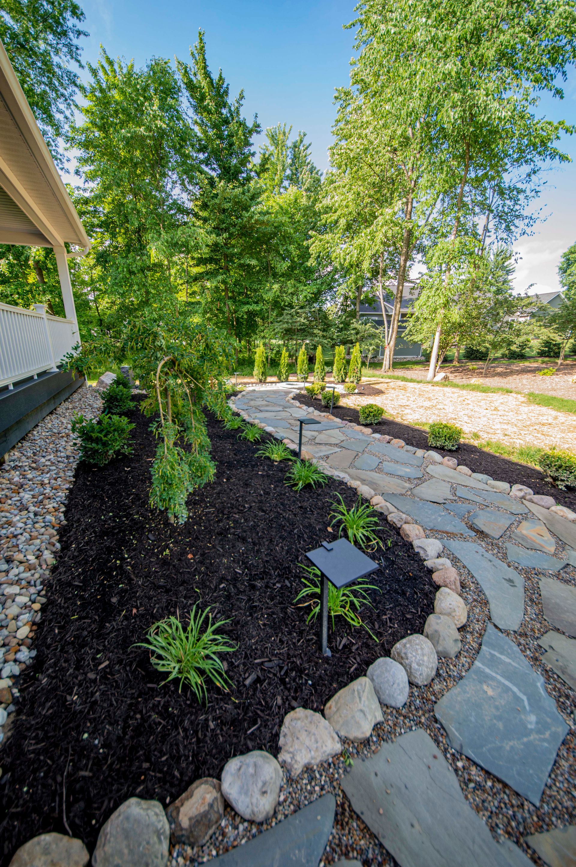 Curved stone path through a landscaped garden with black mulch, rocks, and green trees under a blue sky
