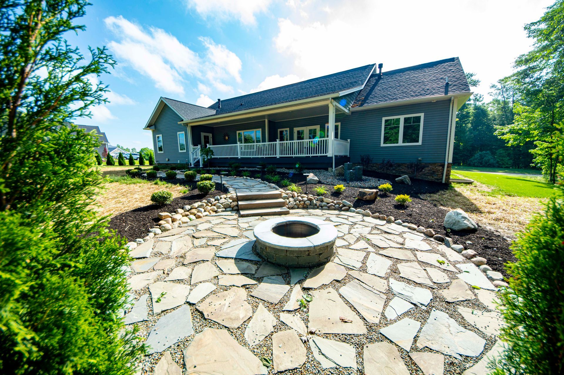 Stone patio with circular fire pit in front of a gray house and porch, surrounded by trees and lawn