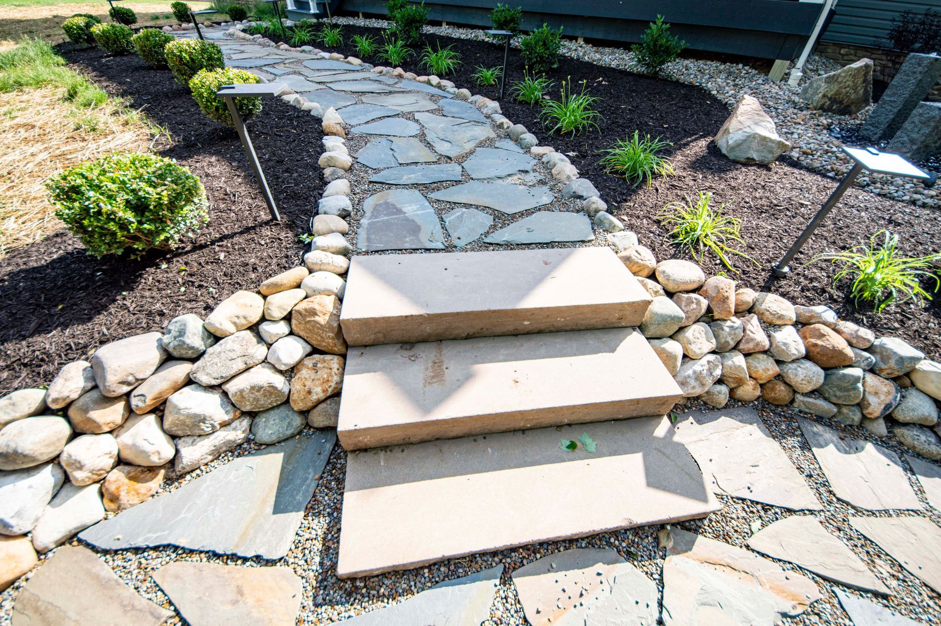 Stone steps in a landscaped garden leading up a flagstone path with mulch and rocks on both sides
