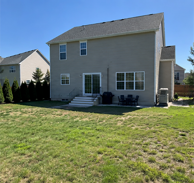 Suburban backyard with a two-story beige house, patio steps, and a grassy lawn under a clear blue sky