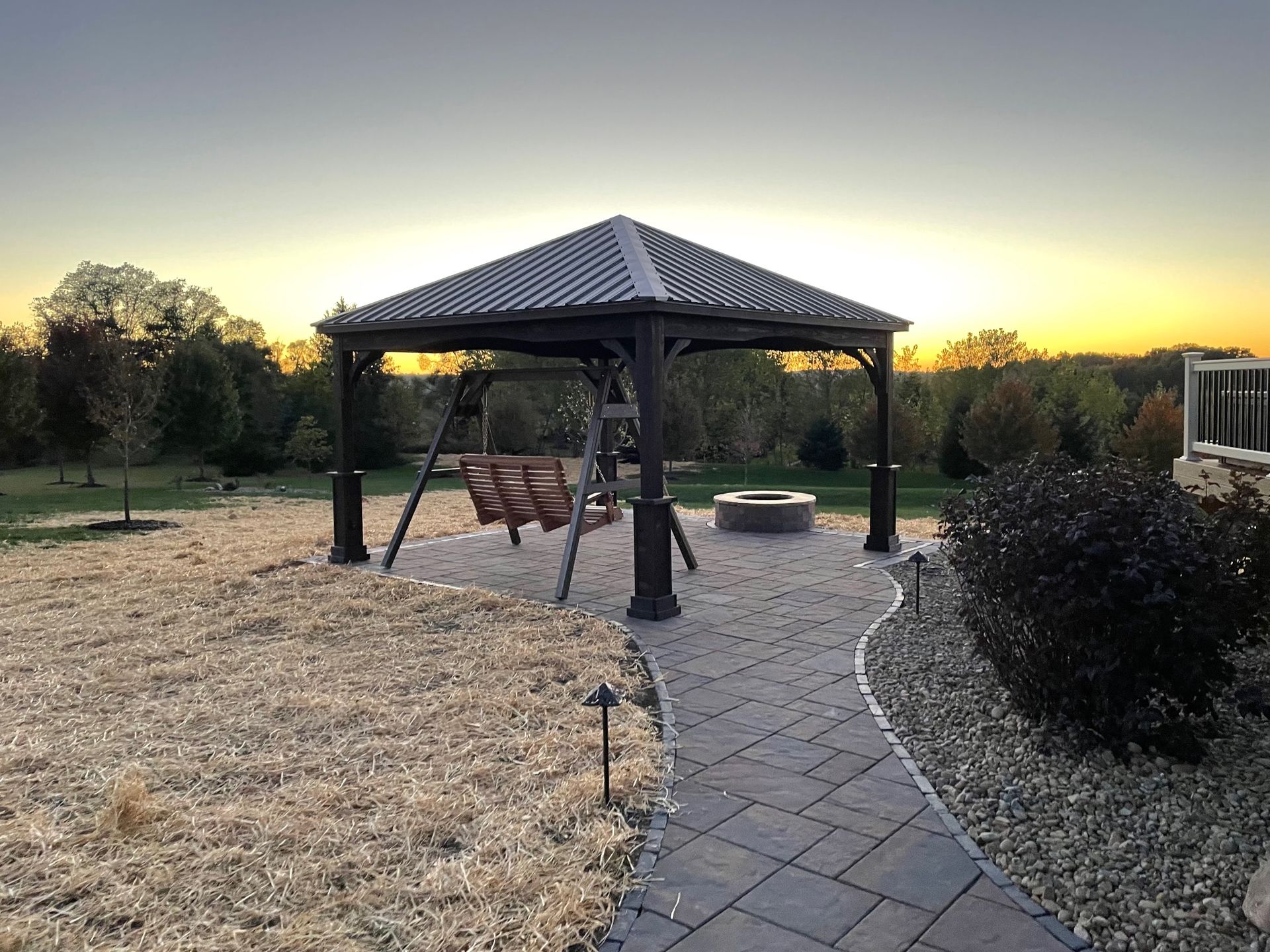 Backyard gazebo at sunset with paved path, fire pit, and dry grass lawn