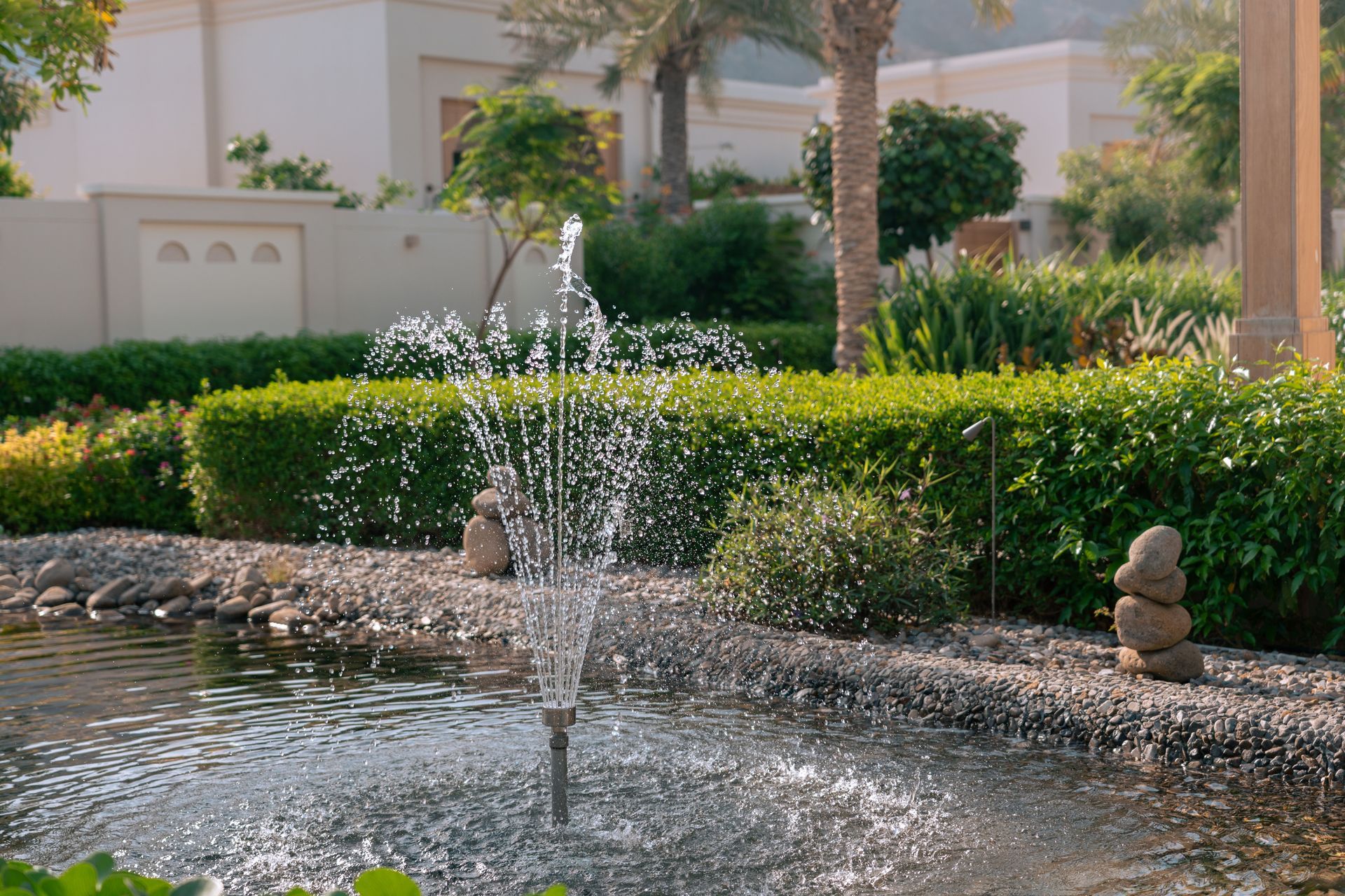Water fountain spraying in a landscaped garden with hedges and a white building in the background