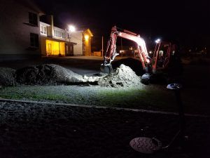 Une excavatrice creuse la nuit près d'un bâtiment ; des lumières vives illuminent la scène.