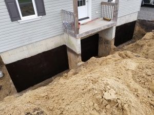 Extérieur d'une maison avec fondations apparentes. Membrane d'étanchéité noire installée, tas de terre devant.