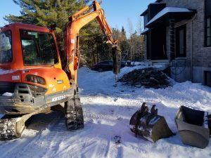 Excavatrice orange, godet et neige un jour d'hiver près d'un bâtiment.