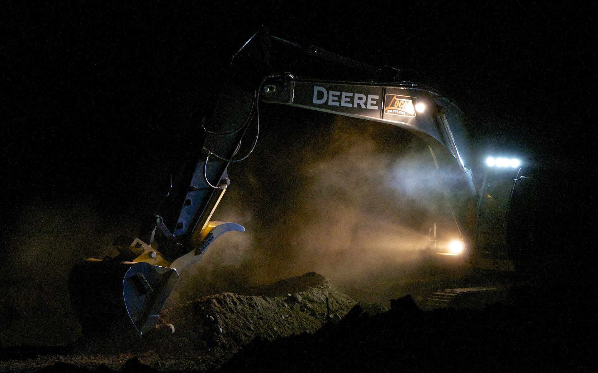 Une excavatrice John Deere creusant la nuit, éclairée par des phares et des phares de travail.