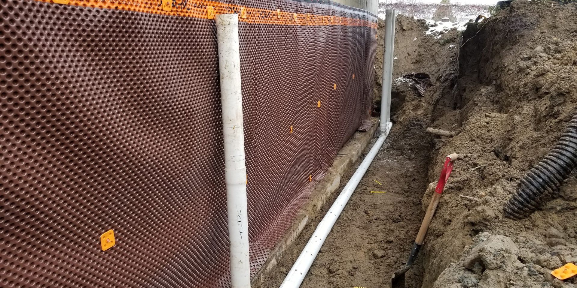 Mur de sous-sol avec membrane alvéolée et tuyau de drainage dans une tranchée. Sol et matériaux de construction visibles.