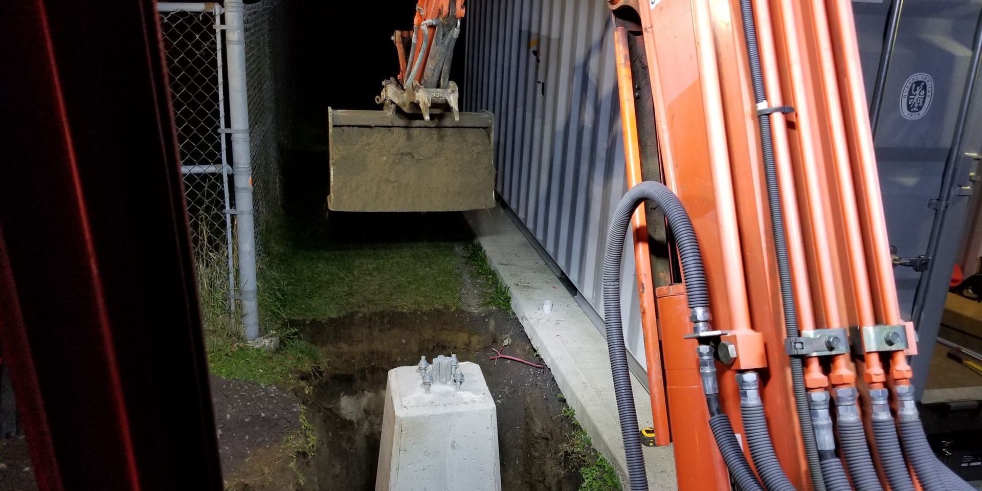 Un godet d'excavatrice déversant de la terre près d'une base en béton, à côté d'une clôture et d'un bâtiment.