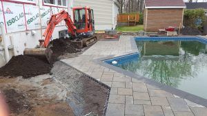 Mini-pelle creusant près d'une maison et d'une piscine, tas de terre, pavés, eau de piscine bleue.