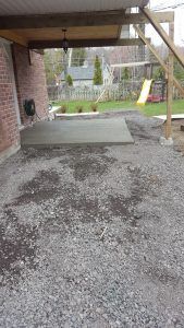 Patio en construction avec base de gravier, mur de briques, pergola en bois et arrière-cour en arrière-plan.