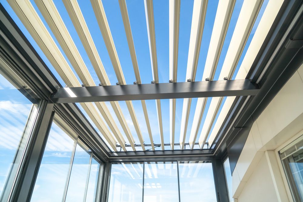 Patio coperto con listelli scuri in metallo della pergola e tende beige retrattili, contro un cielo luminoso