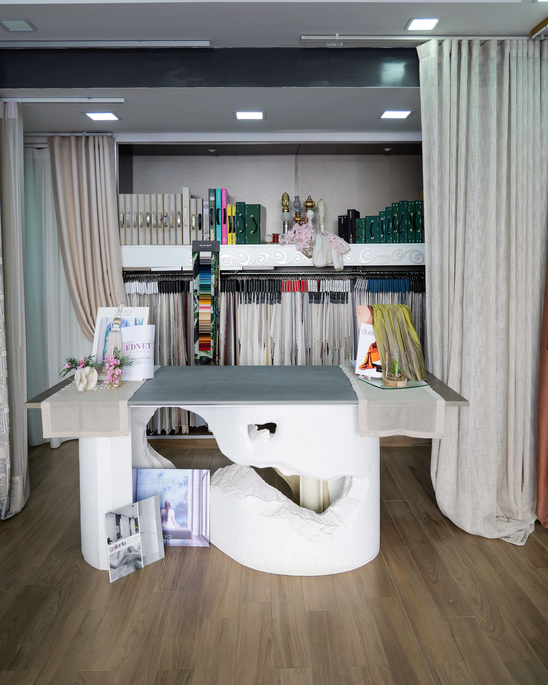 Una scrivania bianca in uno studio domestico, con scaffali pieni di libri e oggetti decorativi, incorniciata da tende a tutta altezza.