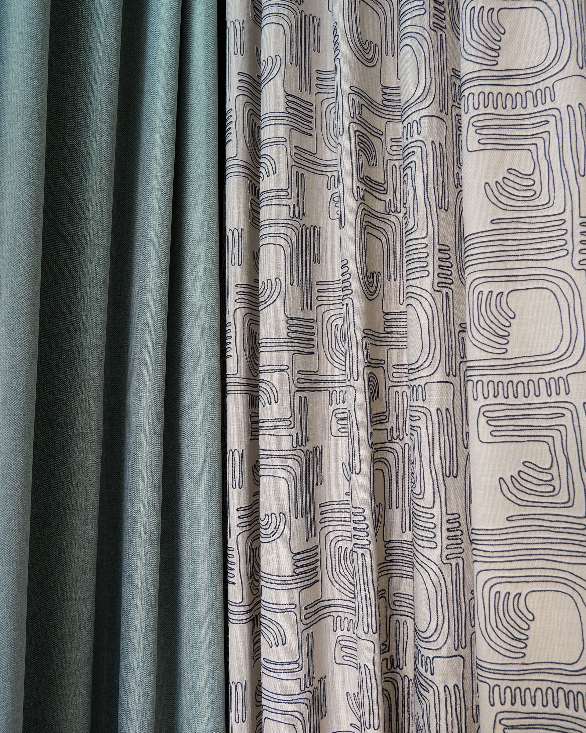 Tende con pannelli di tessuto fantasia blu-verde, beige e grigio appesi uno accanto all’altro