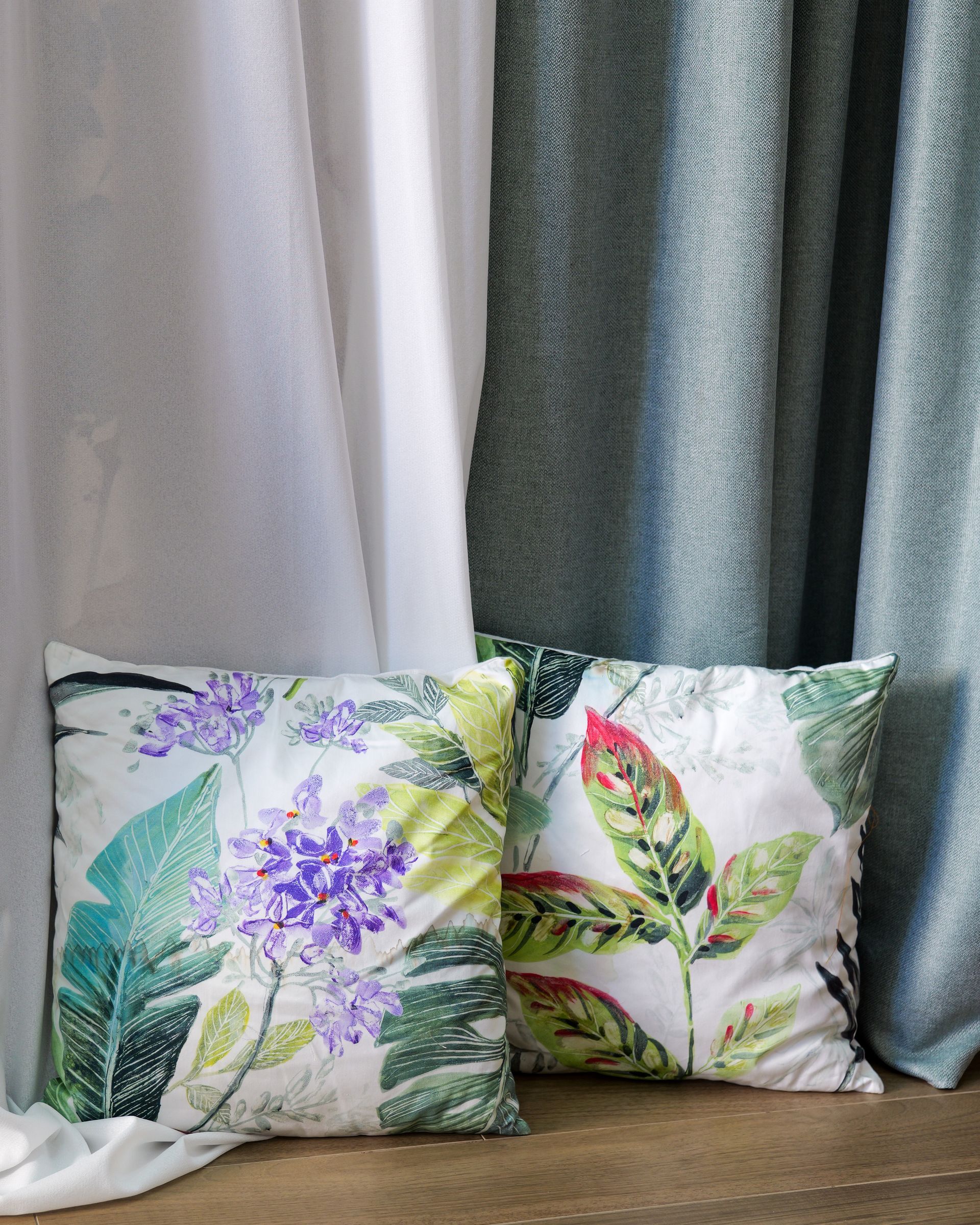 Due cuscini decorativi floreali davanti a tende argentate e grigie, con stampe di foglie verdi e fiori viola