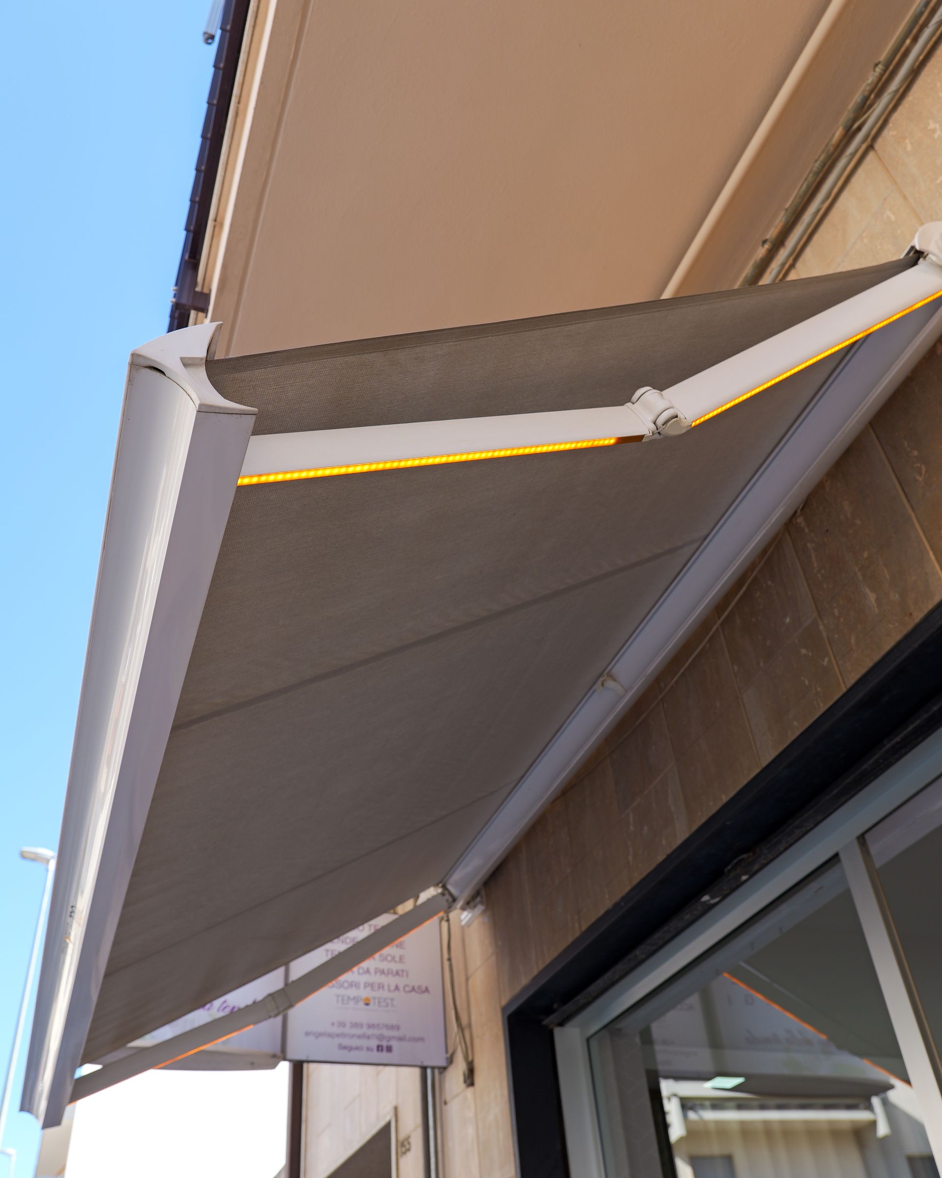 Tenda grigia estesa sopra il passaggio di fronte a un negozio, accanto a una finestra e a una parete beige.