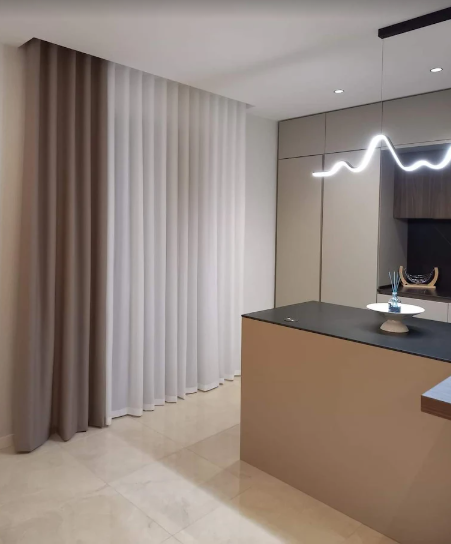 Cucina moderna e minimalista con isola beige, piano di lavoro scuro e tende a tutta altezza accanto