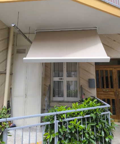 Facciata beige con tenda da sole bianca, finestra e fioriera con piante verdi sulla ringhiera.