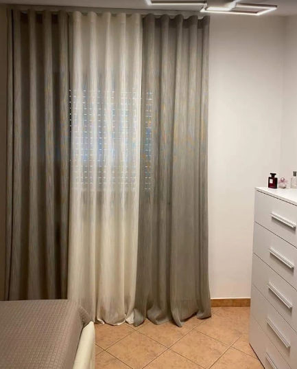 Camera da letto con tende grigie, un bordo del letto e un comò bianco su un pavimento piastrellato color beige.