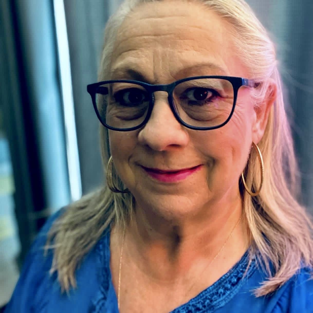 Smiling woman in glasses seated in an office chair, wearing a light denim shirt and orange top.