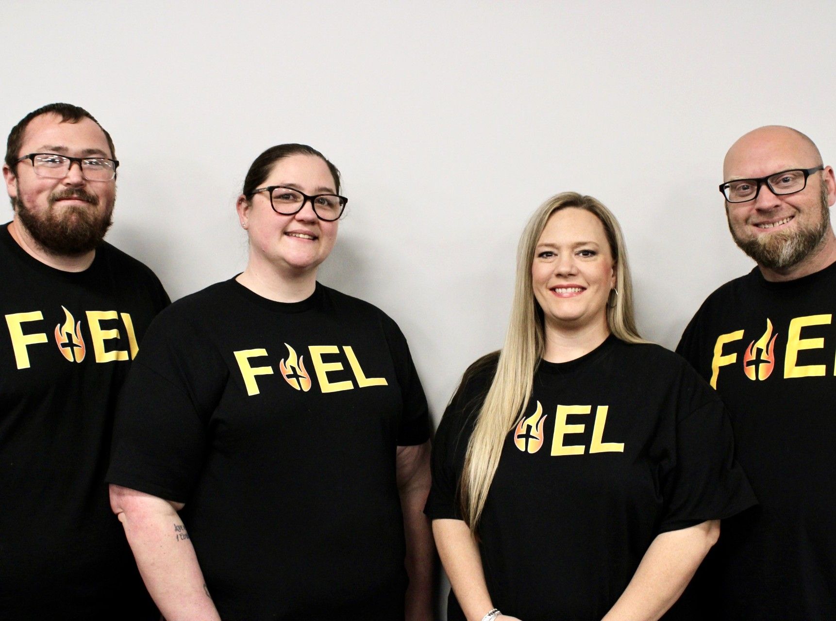 Four people in black FEEL shirts standing against a plain white wall, smiling at the camera.