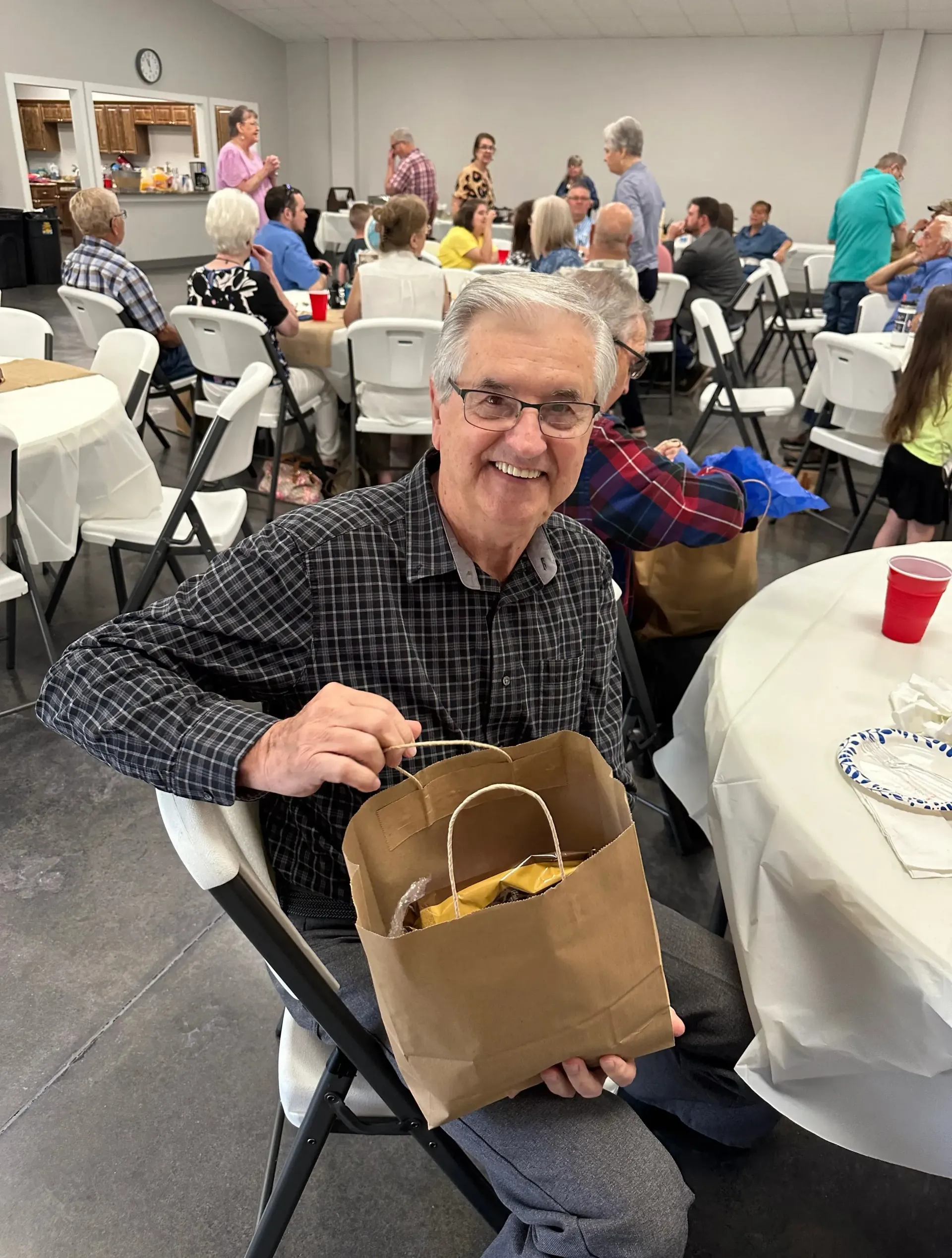 Smiling man holding a paper bag at a community gathering with tables and people in the background
