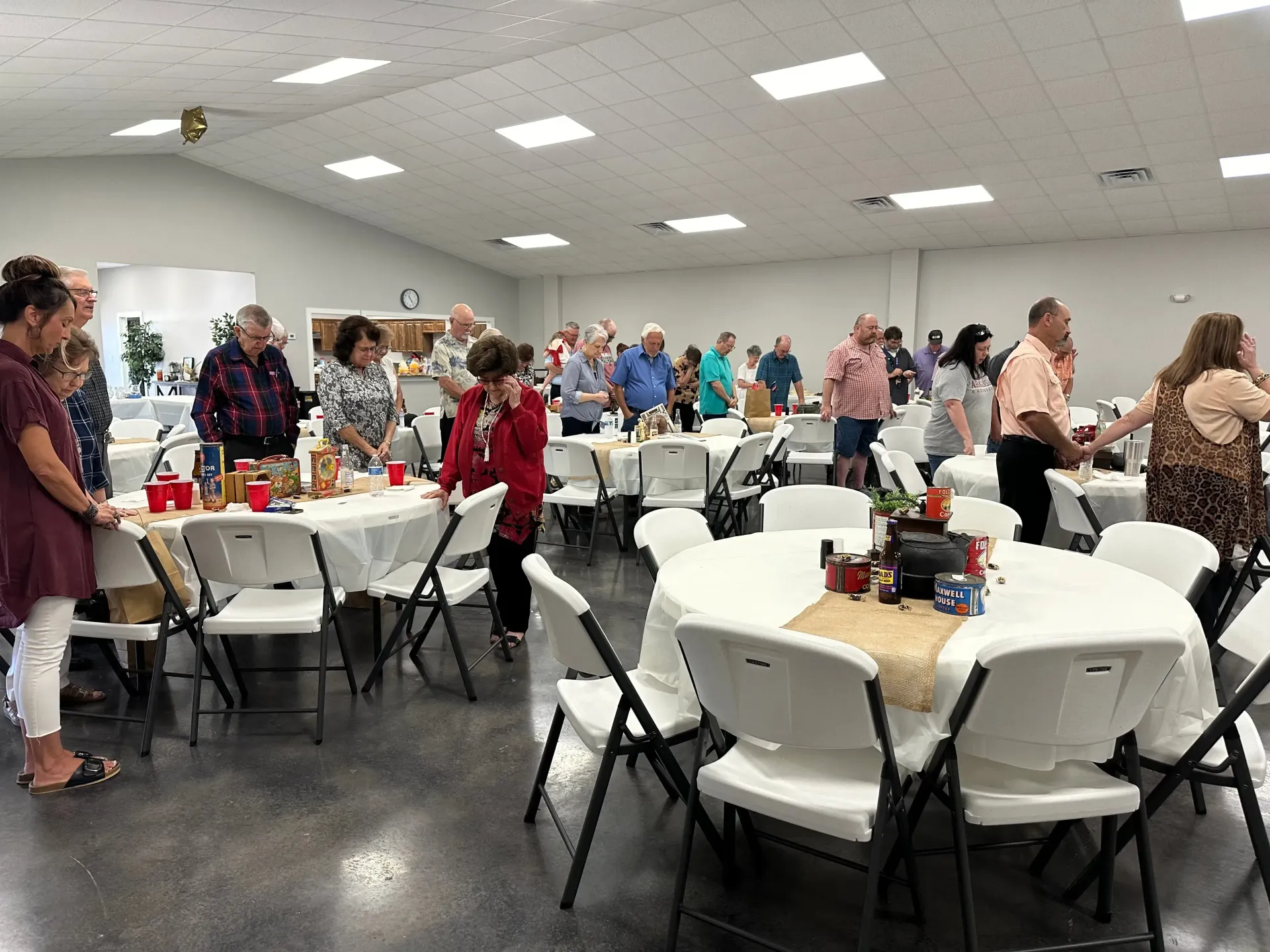 People standing and chatting in a community hall with round tables and white folding chairs
