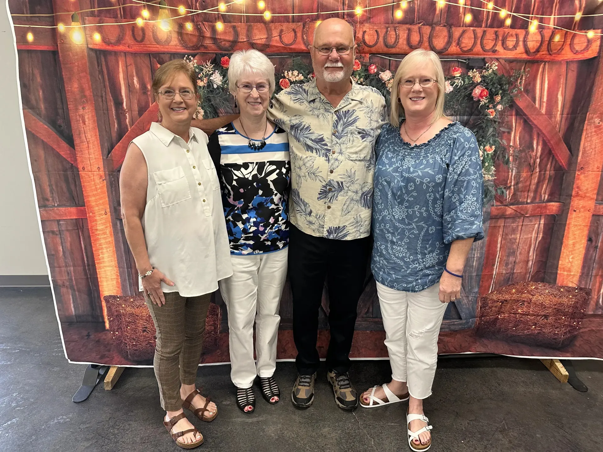Four people standing and smiling in front of a rustic wooden backdrop with string lights