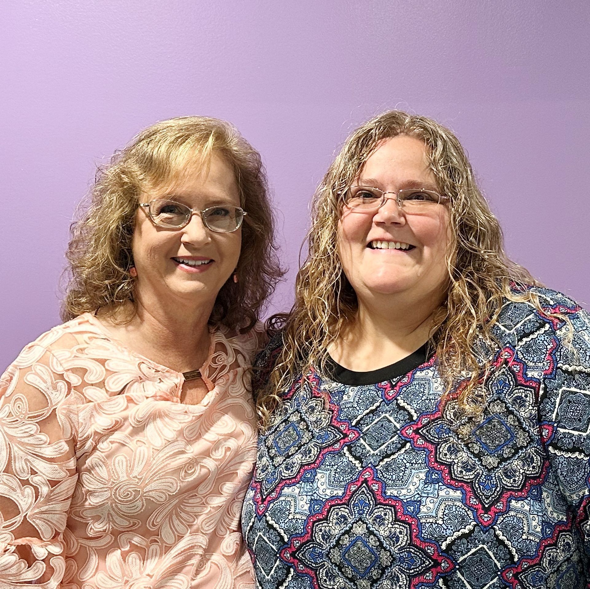 Two smiling people standing together against a purple wall, wearing patterned tops.