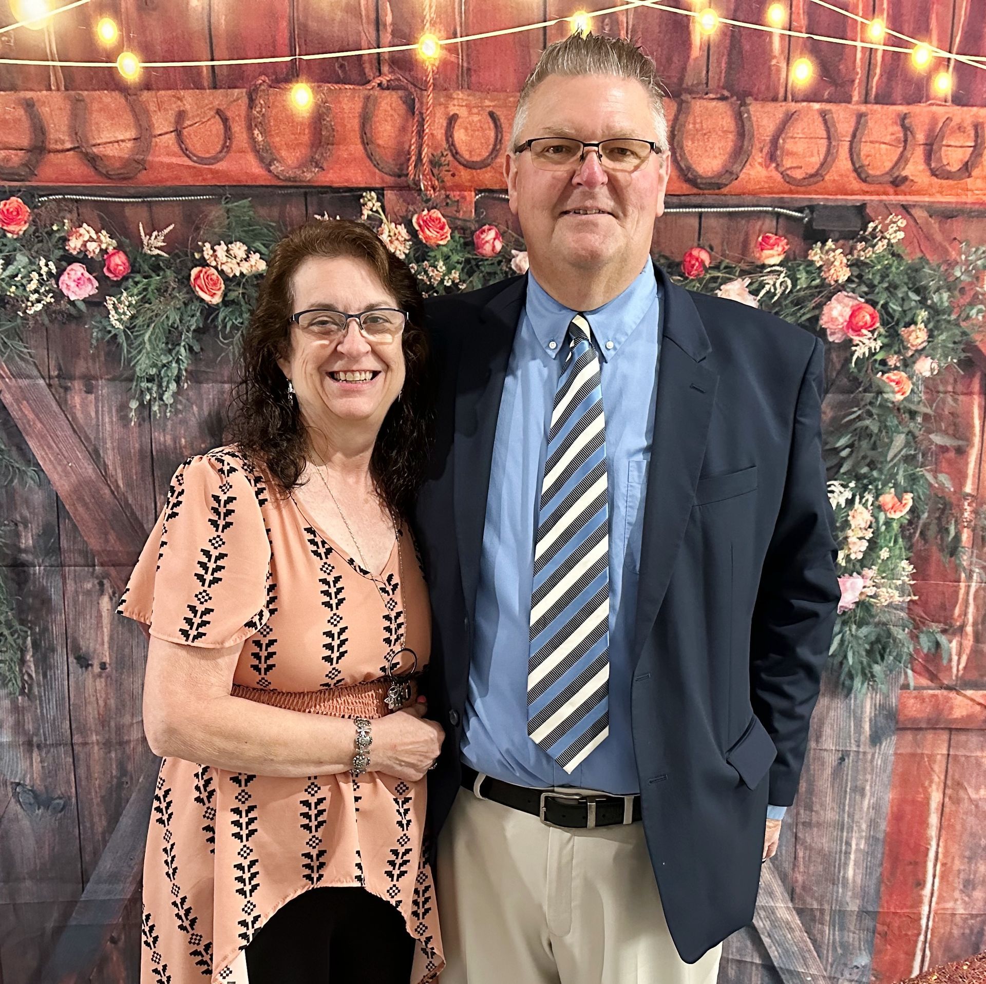 Two people posing in front of a floral backdrop, smiling in formal attire.