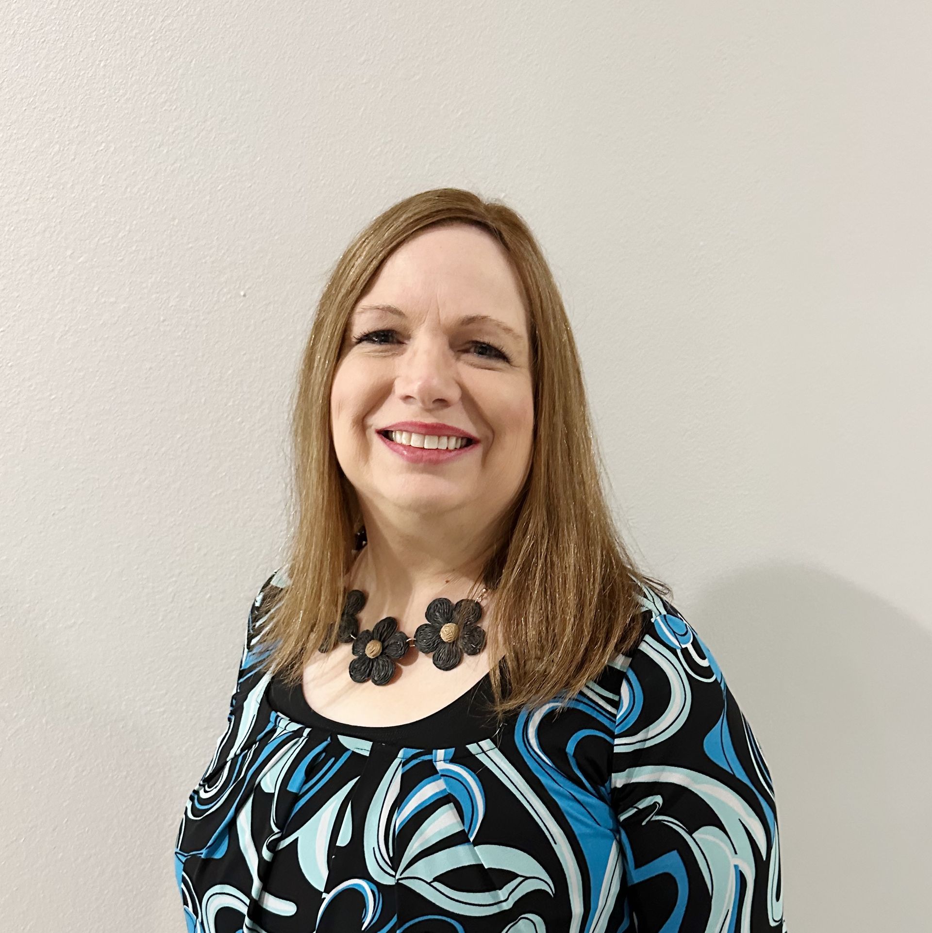 Smiling woman in a black and blue patterned dress and necklace against a plain light wall
