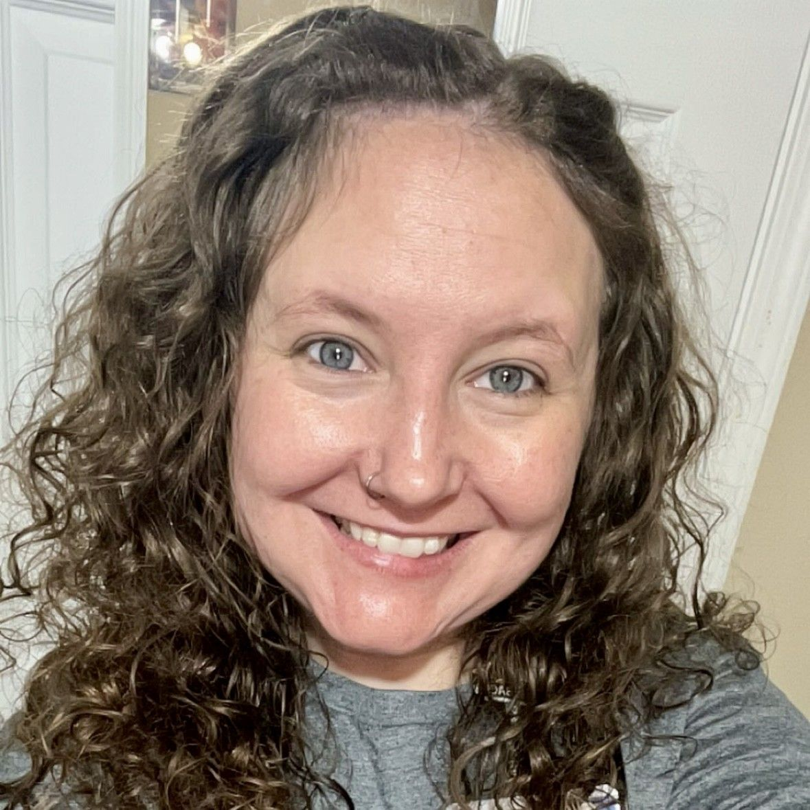 Smiling woman with curly hair in a gray shirt, taking a selfie indoors.