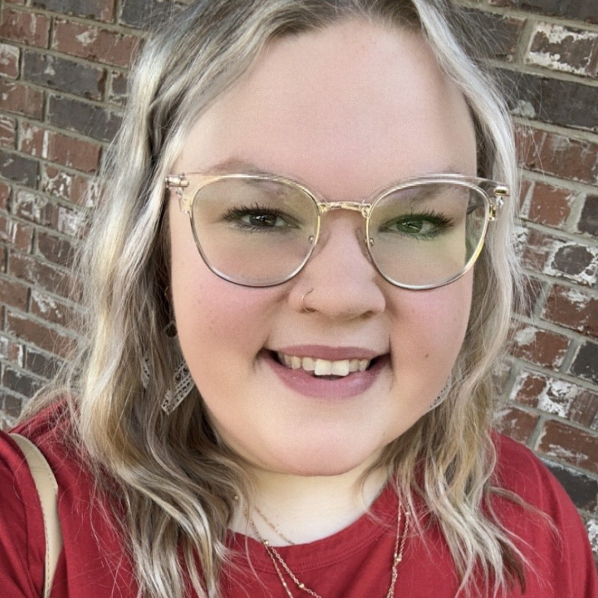 Smiling person with blonde hair and glasses wearing a red shirt in front of a brick wall