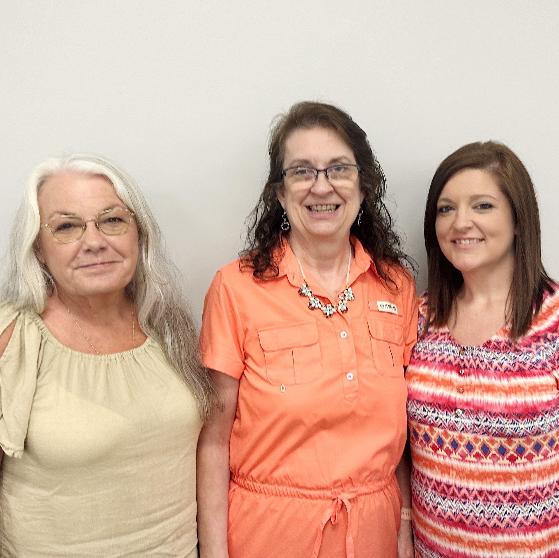 Three people standing together, smiling, against a plain light wall