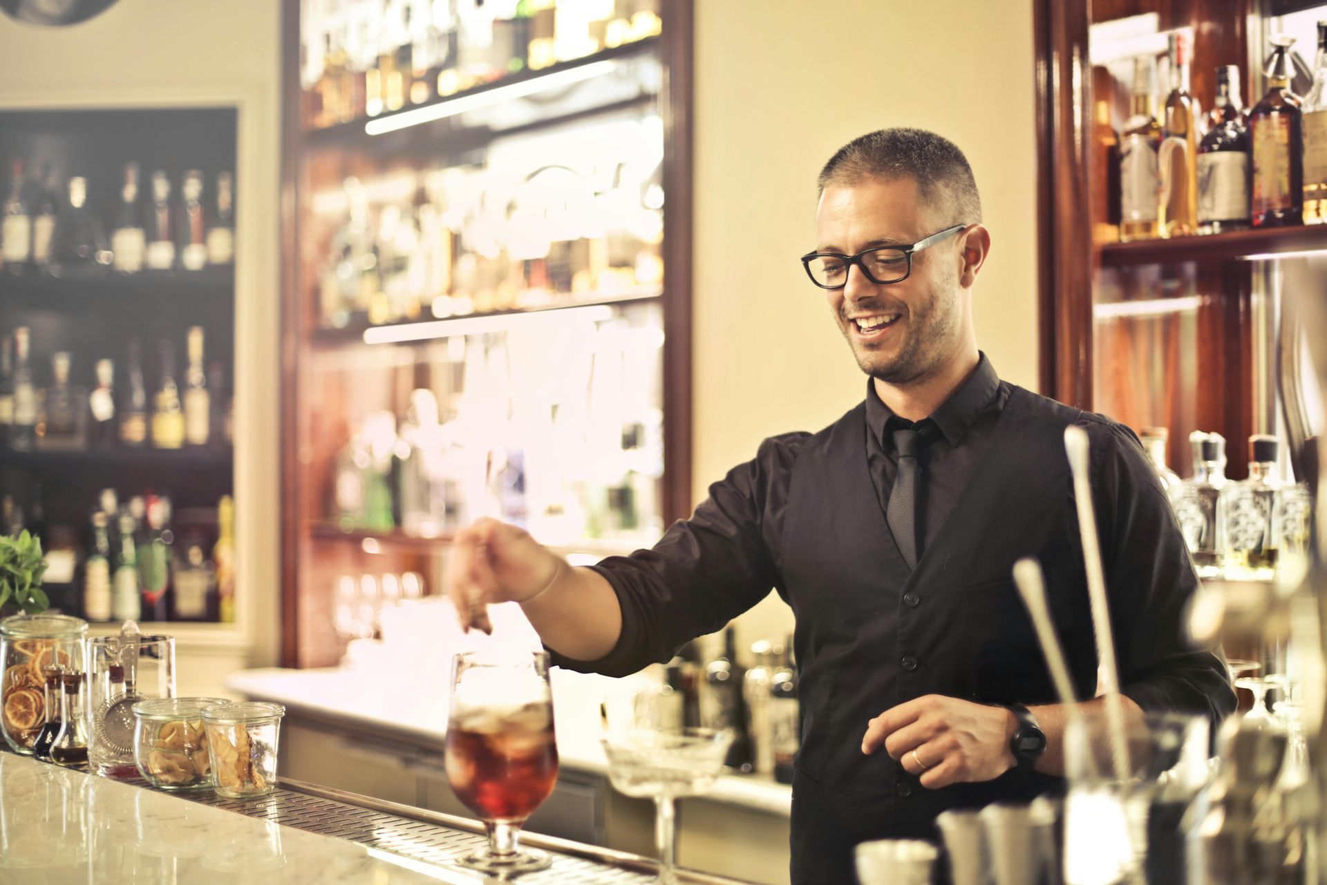 Bartender smiles while stirring a red cocktail behind a bar with bottles.