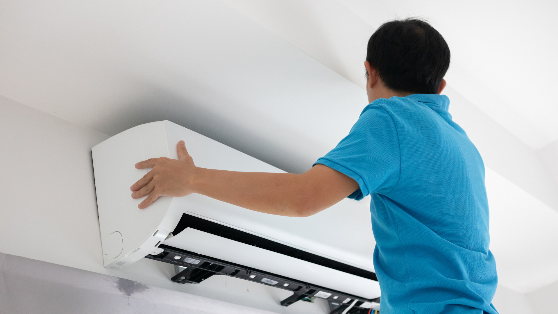 Person installing a white wall-mounted air conditioner indoors, wearing a blue shirt