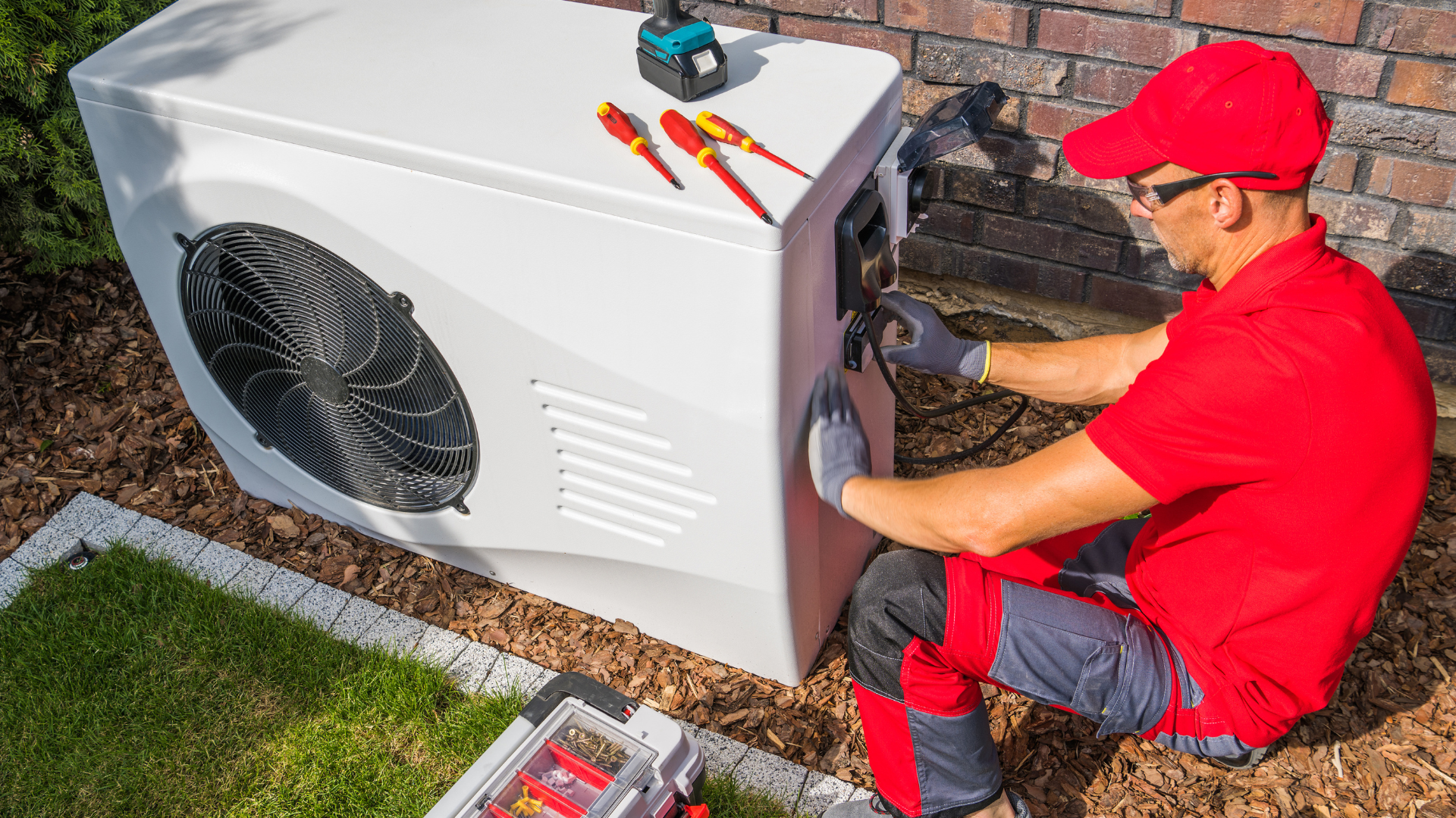 A technician in a red uniform installs an outdoor heat pump unit near a brick wall, with tools placed on top.