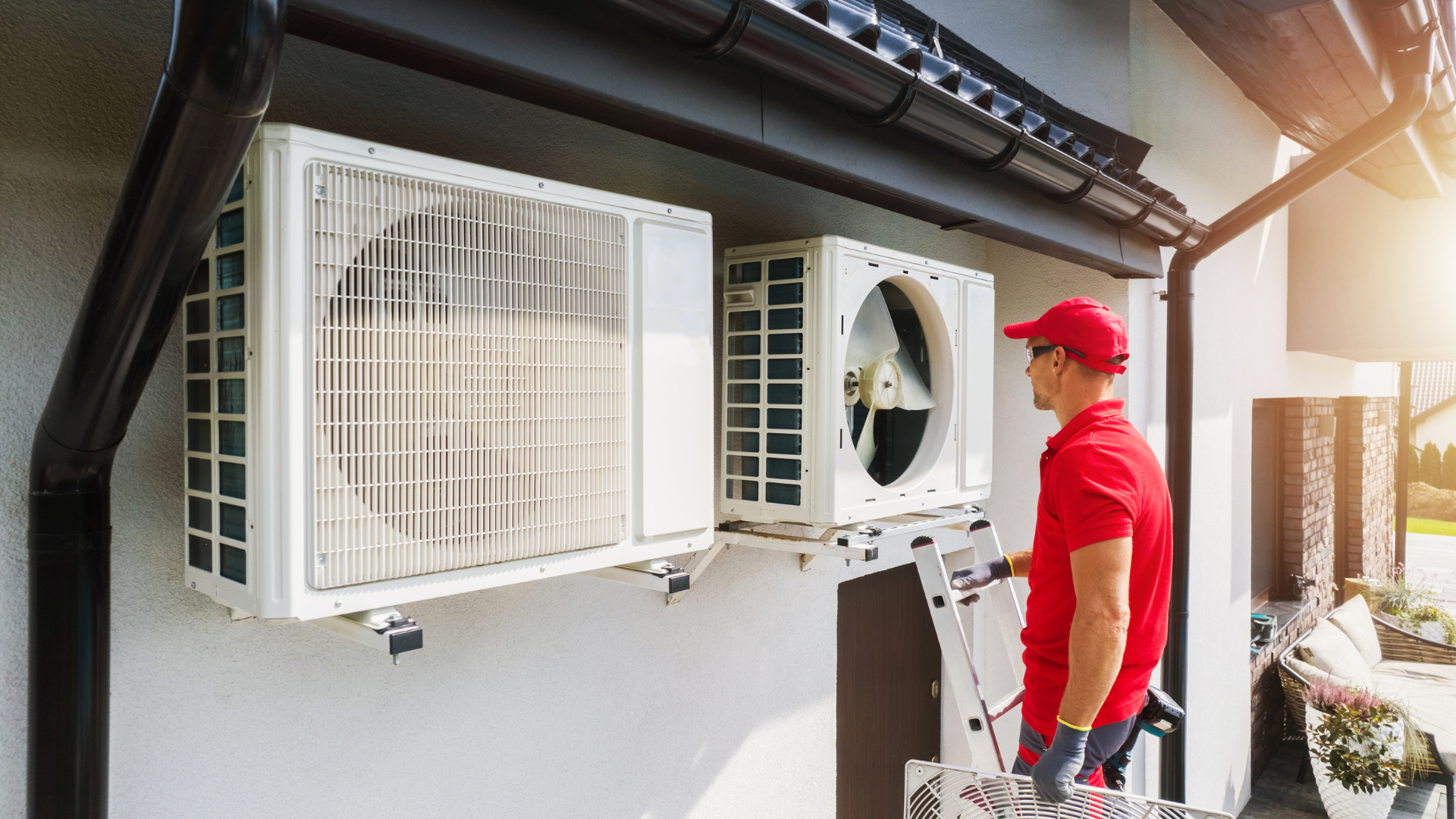 A technician in a red uniform installs or repairs an outdoor air conditioning unit on the side of a house.