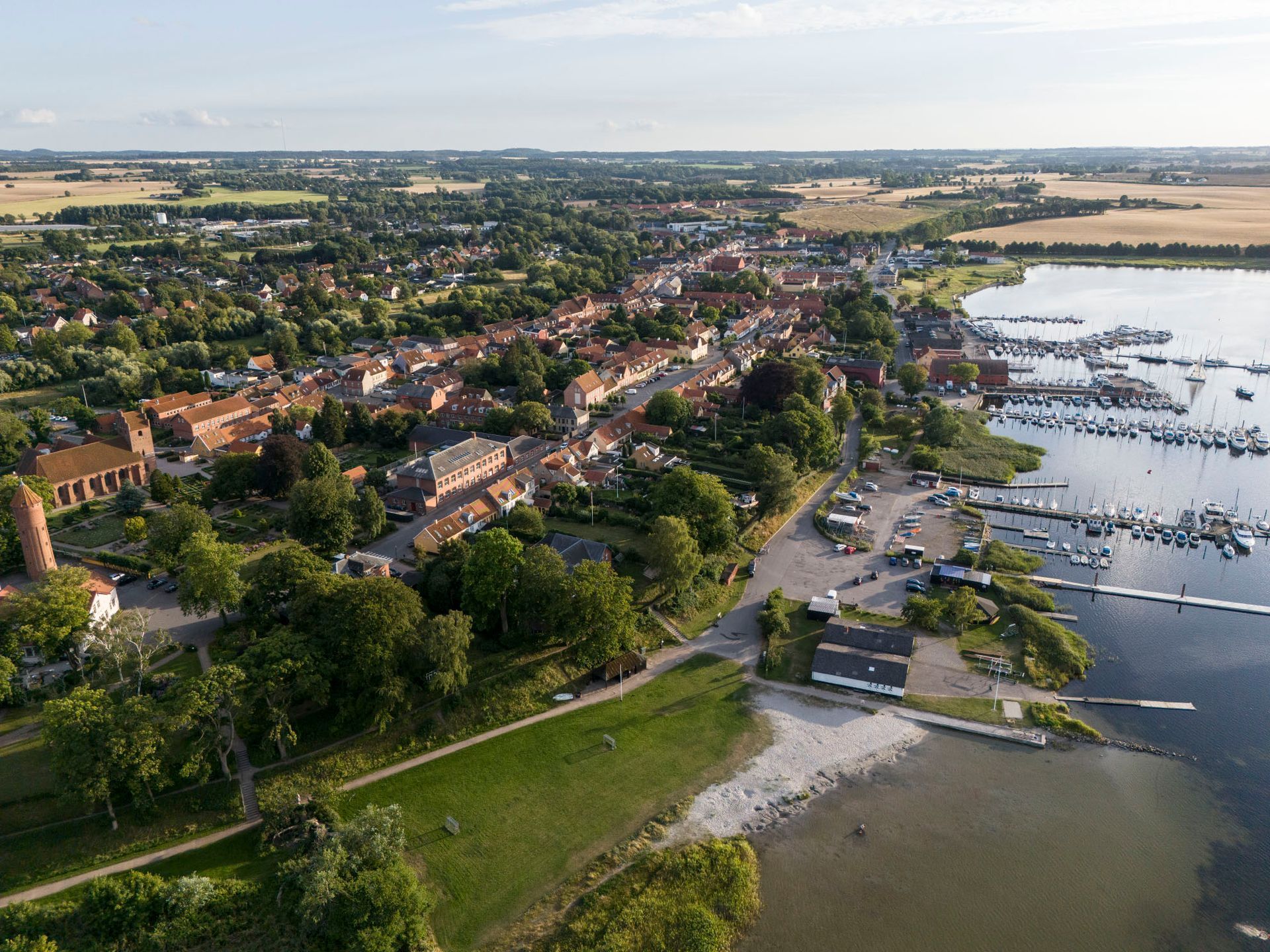 Luftfoto af en by ved en havn, bygninger med røde tage, både fortøjet, grønne træer og marker.