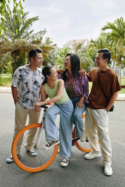 Four friends standing with an orange bicycle on a path, smiling and chatting in a sunny park