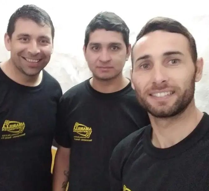 Tres personas sonrientes con camisas negras posando en el interior de una habitación que parece un taller.