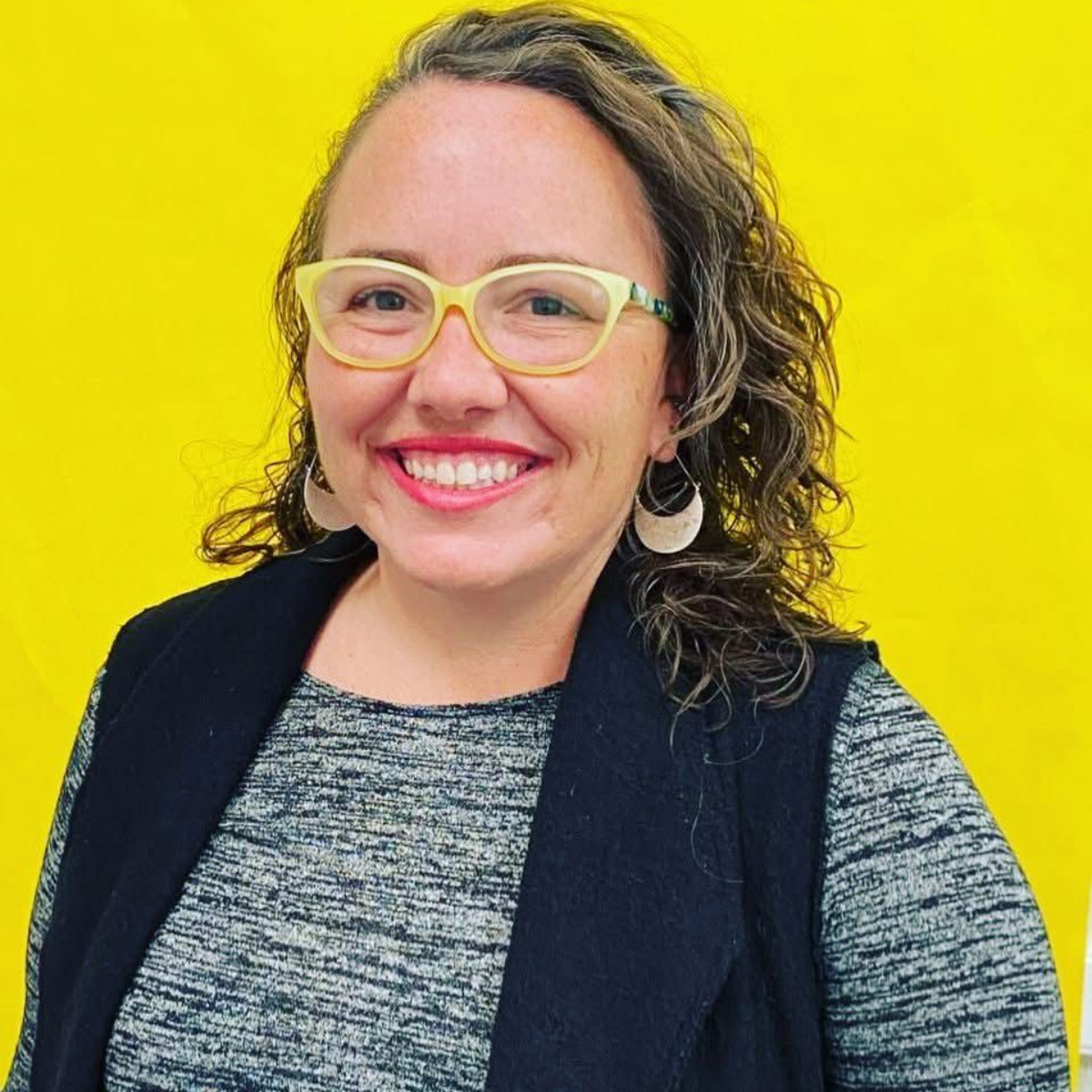 White woman with yellow rimmed glasses and looped earrings. Brown wavy hair with blond highlights--smiling at the camera.