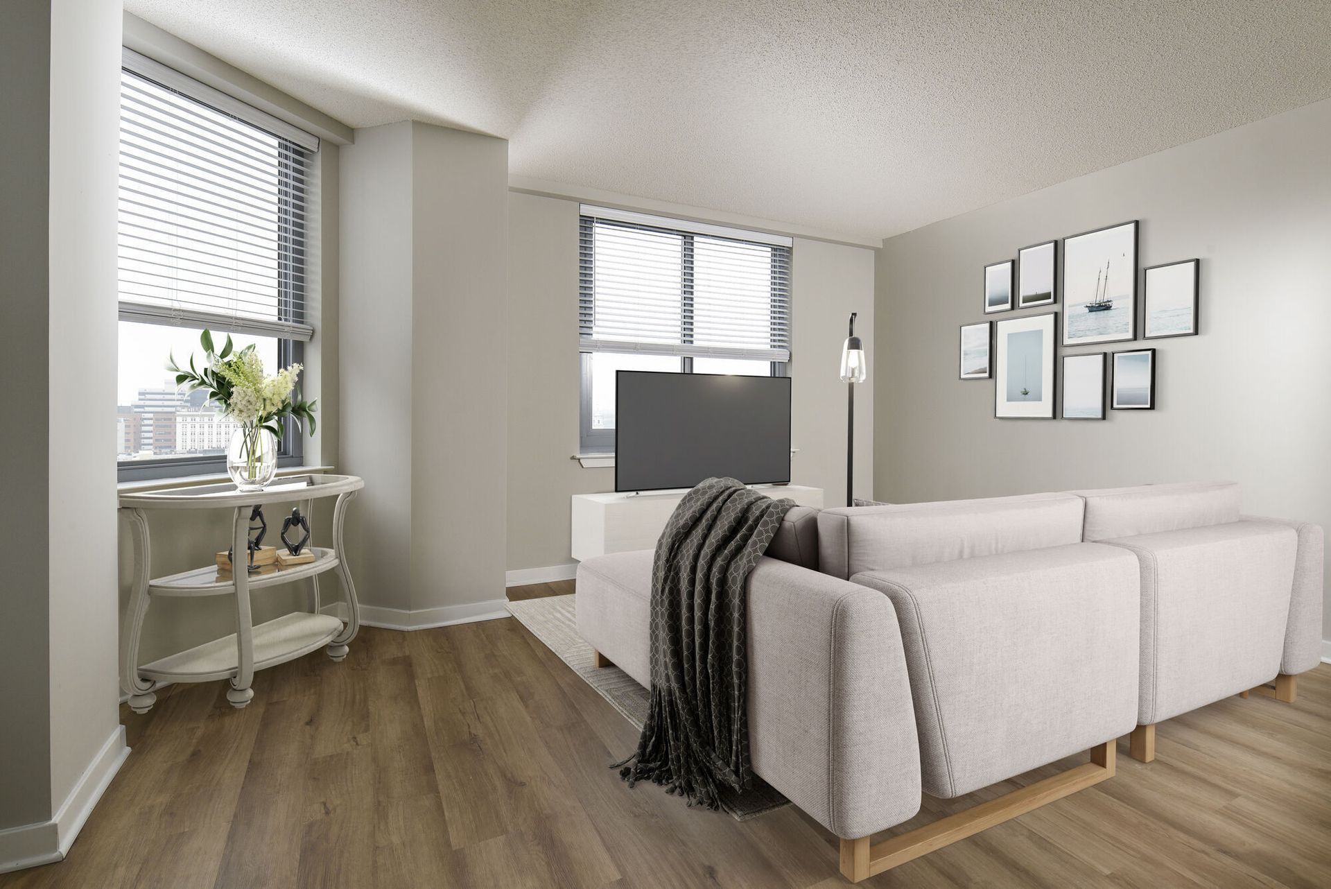 Living room with gray sectional, TV on white console, wood floors, window blinds, and wall art.