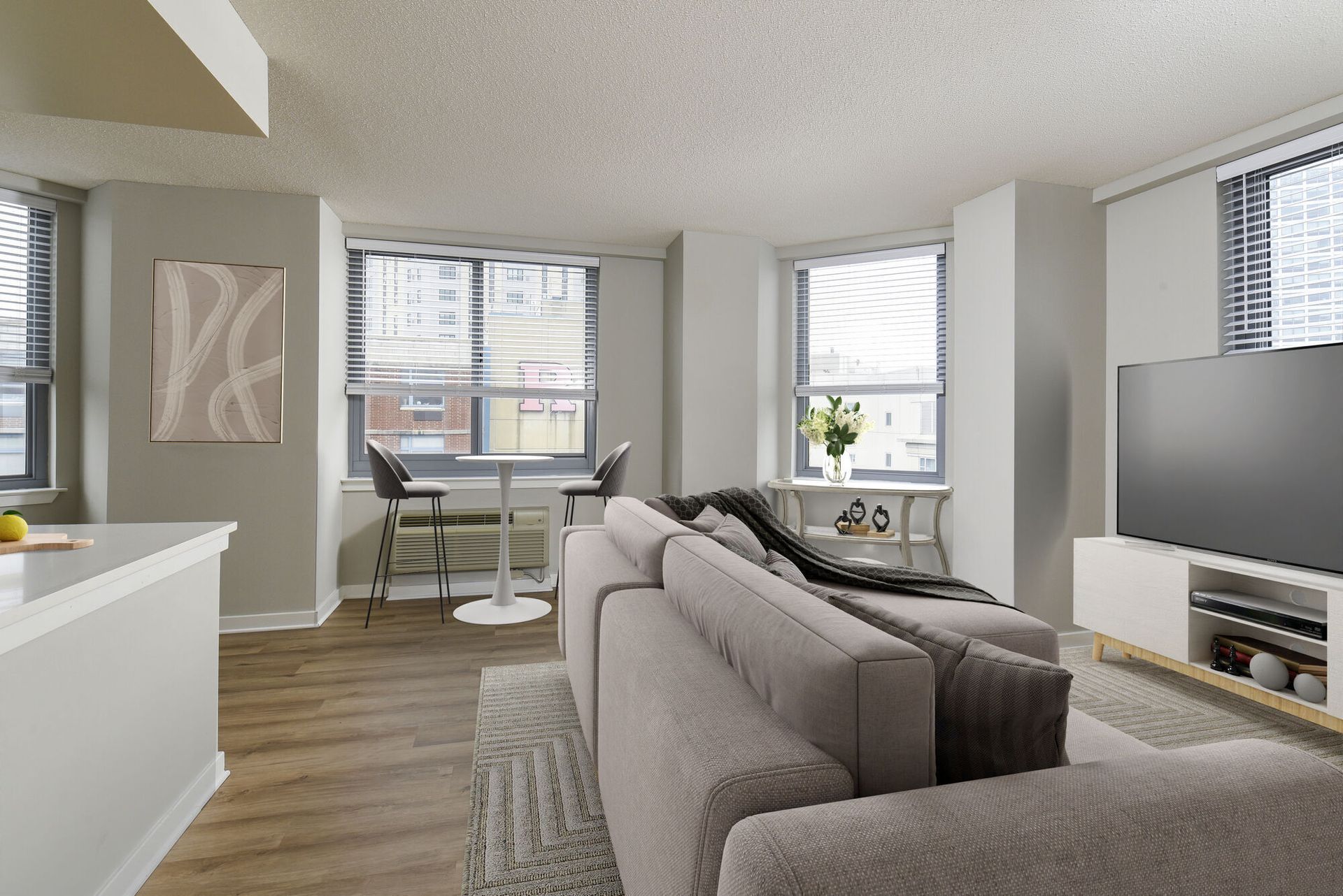 Bright living room with a gray sectional, large windows with blinds, and a TV stand.