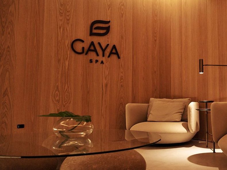 Hall de entrada do spa com painéis de madeira, logotipo do Gaya Spa, cadeira bege e mesa de centro de vidro.