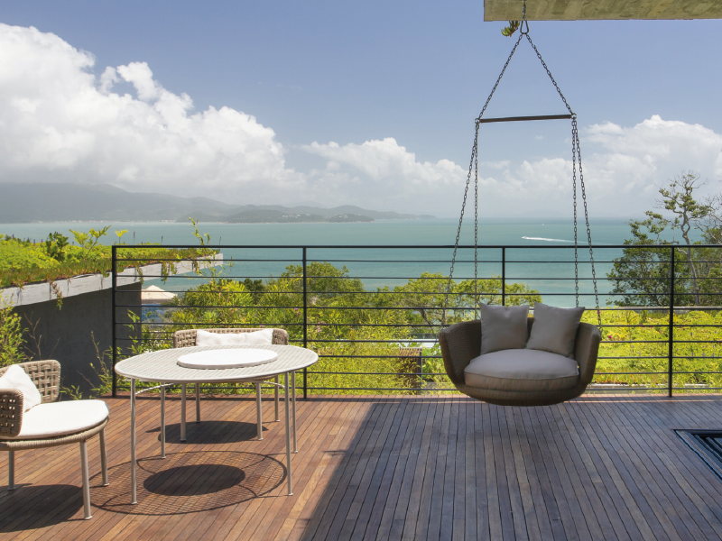Deck moderno com cadeira suspensa, mesa branca e vista para o mar sob um céu parcialmente nublado.