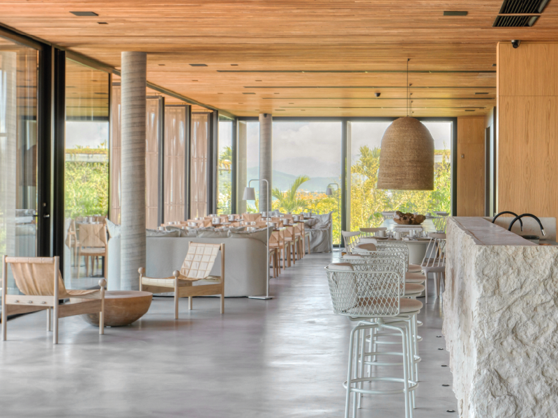 Restaurante moderno e luminoso com teto de madeira, cadeiras brancas e vista para o mar através de grandes janelas.