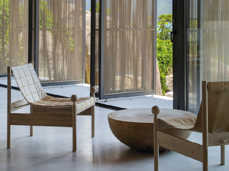 Sala de estar moderna com duas cadeiras de madeira e uma mesa redonda baixa junto a cortinas translúcidas e uma porta aberta.